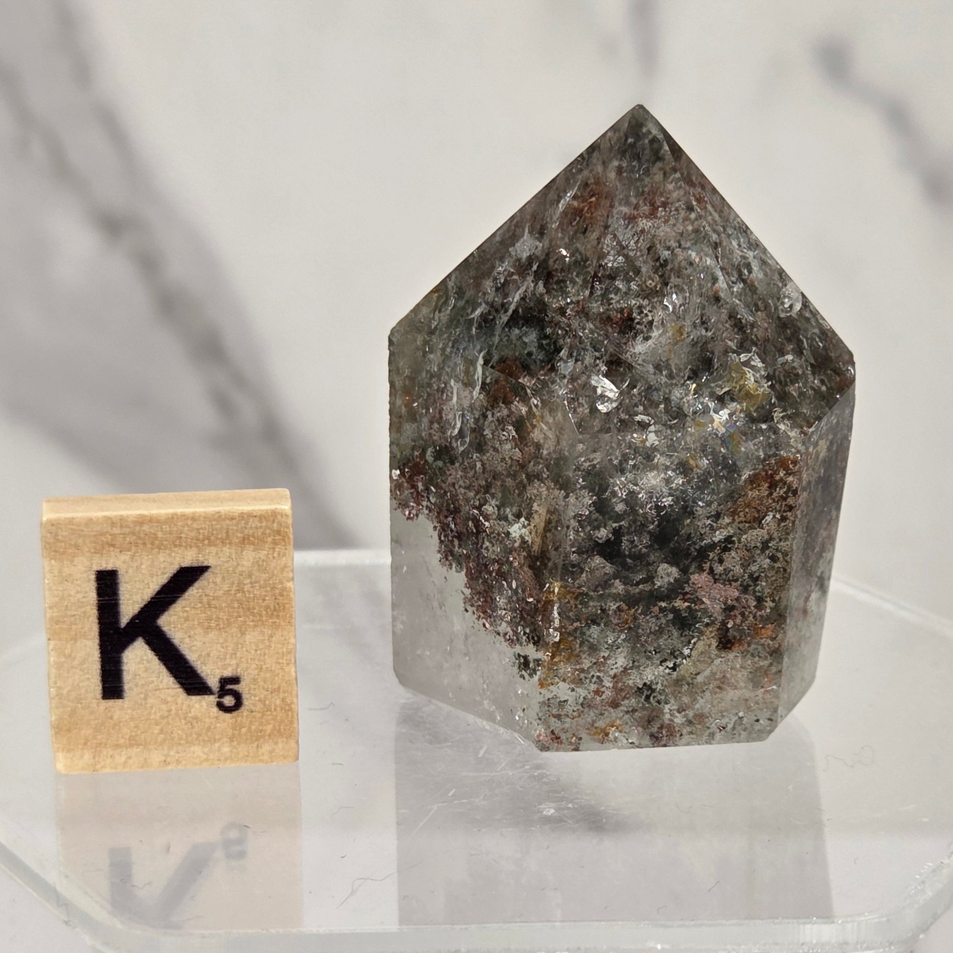 A natural pointed crystal gemstone with a mottled gray, brown, and white speckled pattern, displayed on a clear acrylic stand next to a wooden Scrabble tile spelling 'K' with a 5.