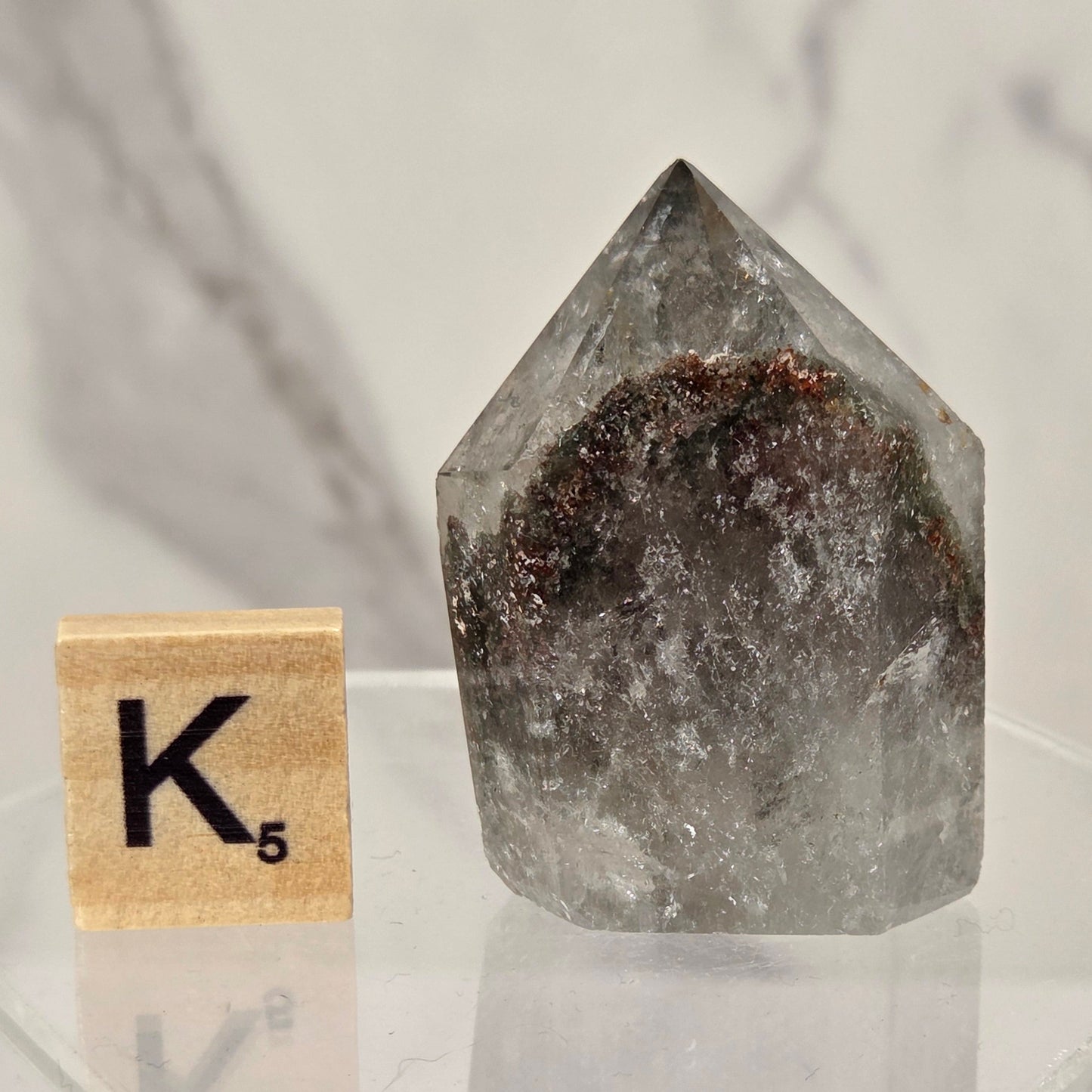 A smoky quartz crystal point with a dark brown interior, displayed on a clear acrylic stand next to a wooden Scrabble tile showing the letter K and value 5, against a white marble background.