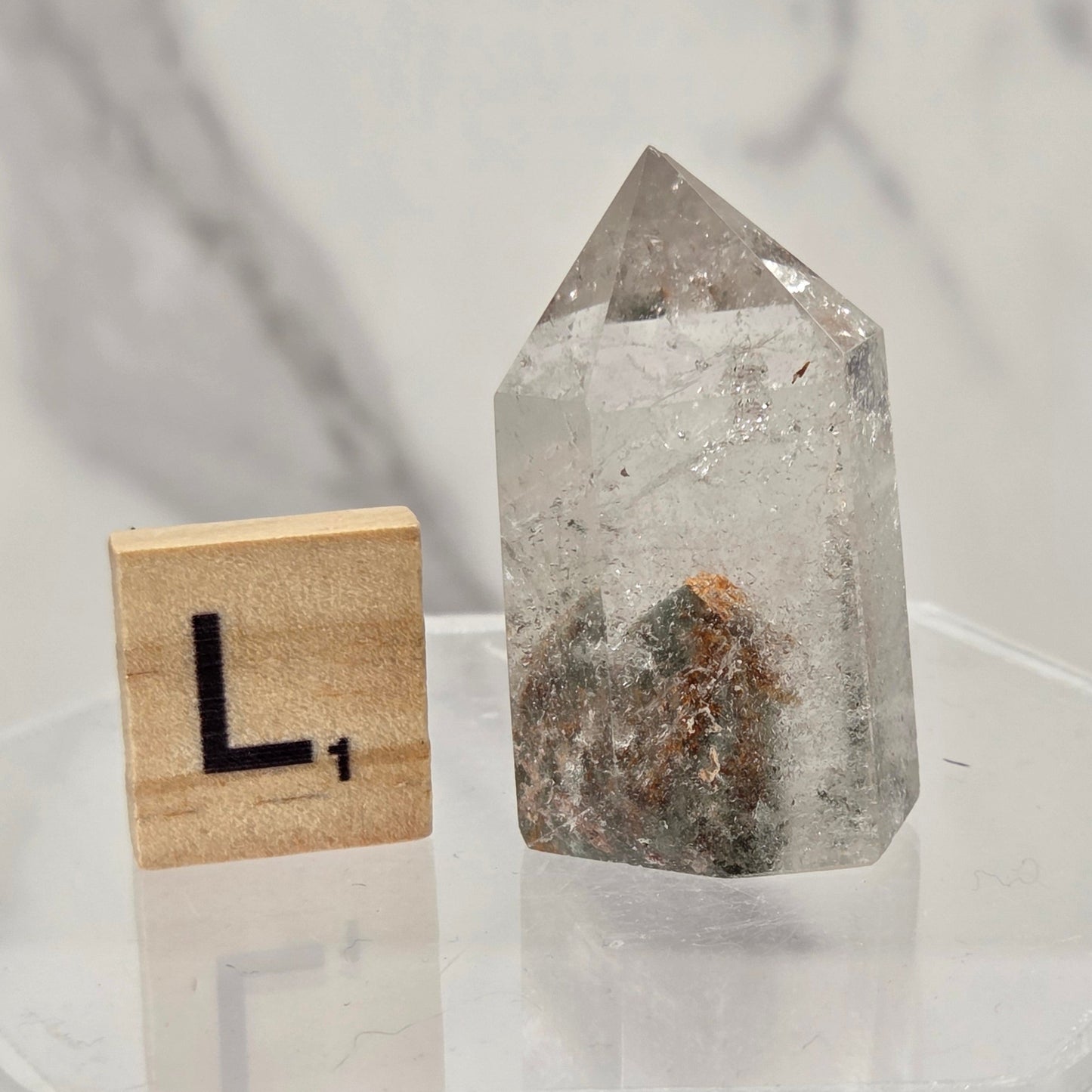 A clear quartz point crystal with internal inclusions, displayed next to a wooden Scrabble tile on a clear acrylic stand against a marble-patterned background.