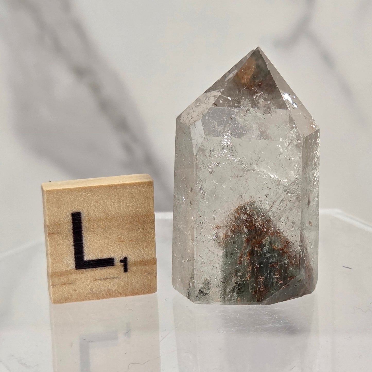 A clear quartz crystal point with internal inclusions sits on a clear acrylic display block next to a wooden Scrabble tile showing the letter 'L'.