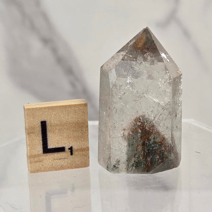 A clear quartz crystal point with internal inclusions sits on a clear acrylic display block next to a wooden Scrabble tile showing the letter 'L'.