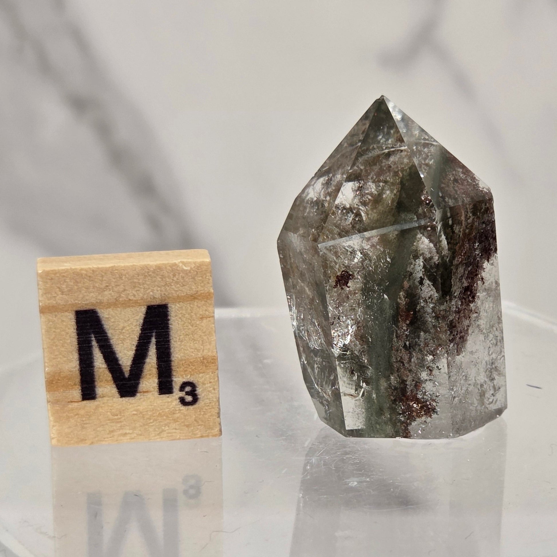 A natural clear quartz point crystal with dark inclusions, displayed next to a wooden Scrabble tile showing the letter M on a clear acrylic stand against a marble-patterned background.