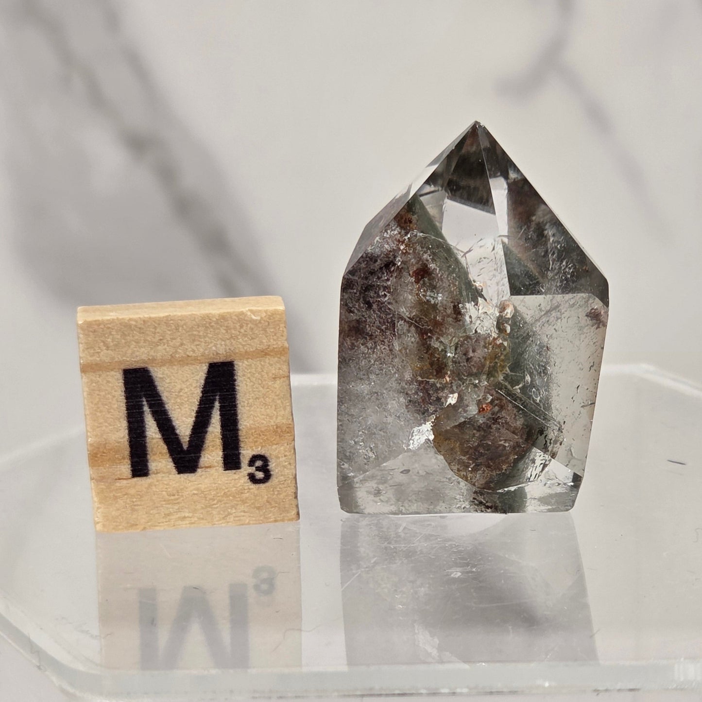 A clear faceted crystal specimen with internal brown and gray inclusions, displayed next to a wooden Scrabble tile marked 'M3' on a clear acrylic stand against a light marble-patterned background.