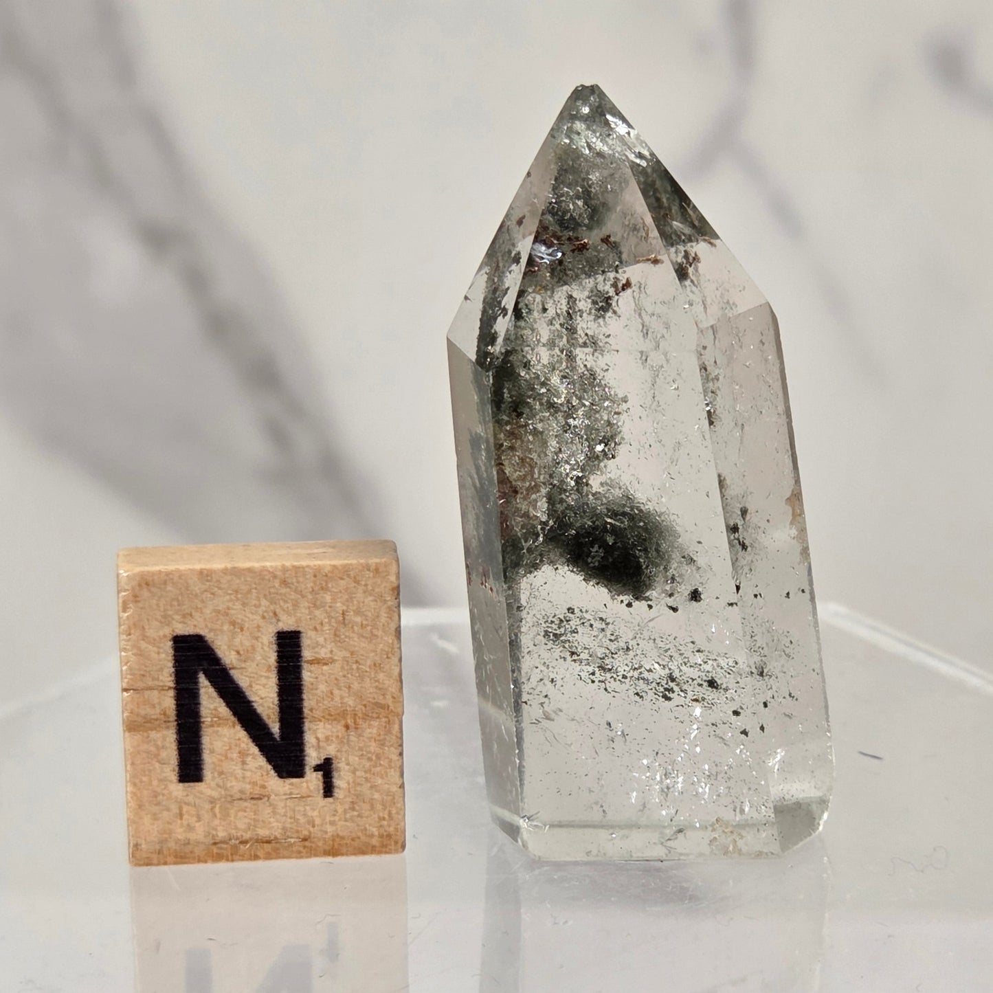 A clear quartz crystal point with internal inclusions sits on a clear acrylic stand next to a wooden Scrabble tile showing the letter 'N'.