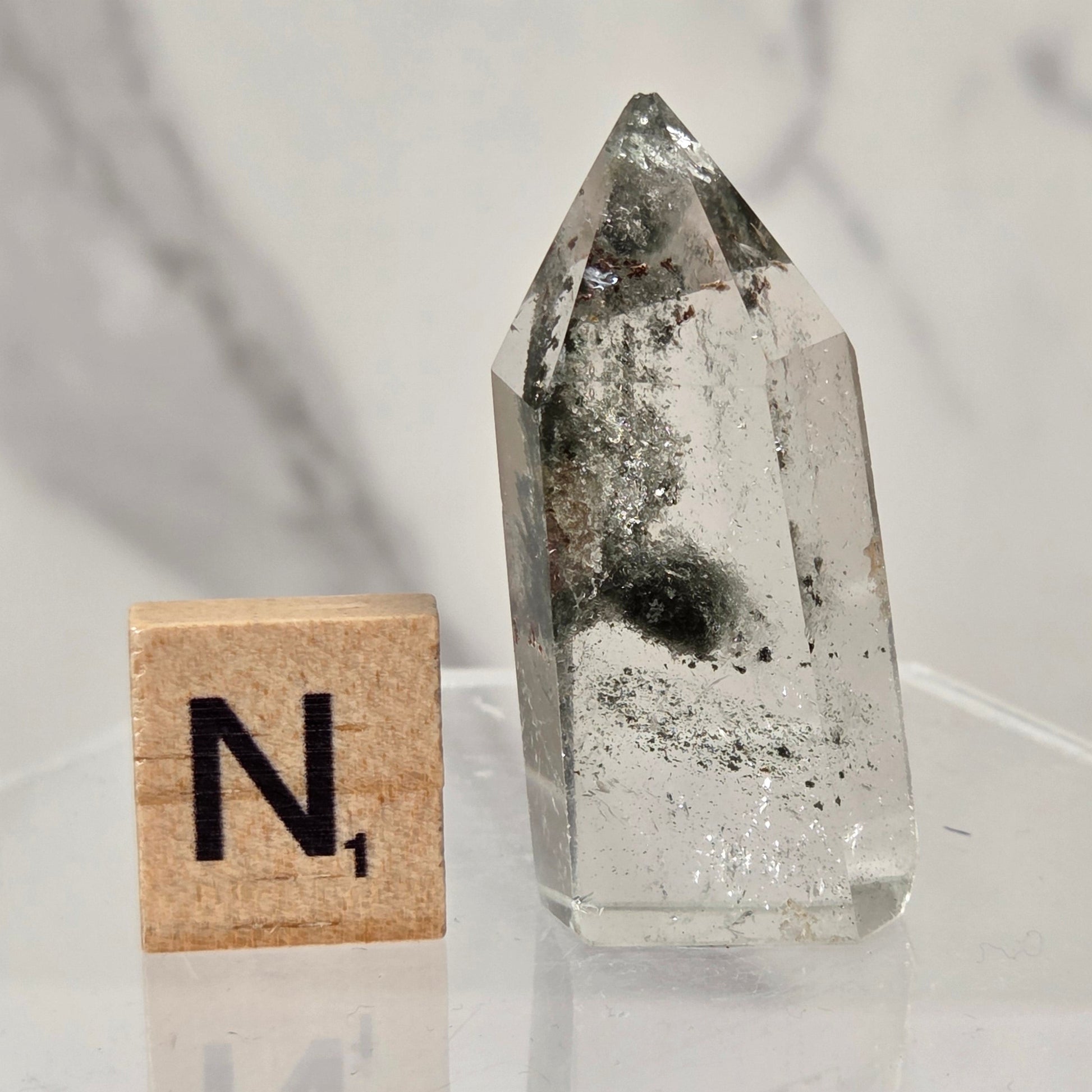 A clear quartz crystal point with internal inclusions sits on a clear acrylic stand next to a wooden Scrabble tile showing the letter 'N'.