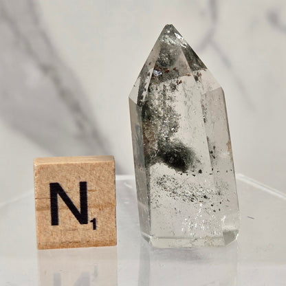 A clear quartz crystal point with internal inclusions sits on a clear acrylic stand next to a wooden Scrabble tile showing the letter 'N'.