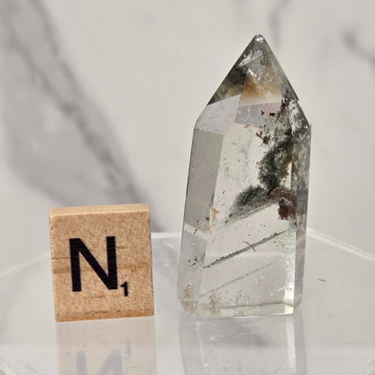 A clear quartz crystal point with internal inclusions sits on a clear acrylic stand next to a wooden Scrabble tile showing the letter 'N'.