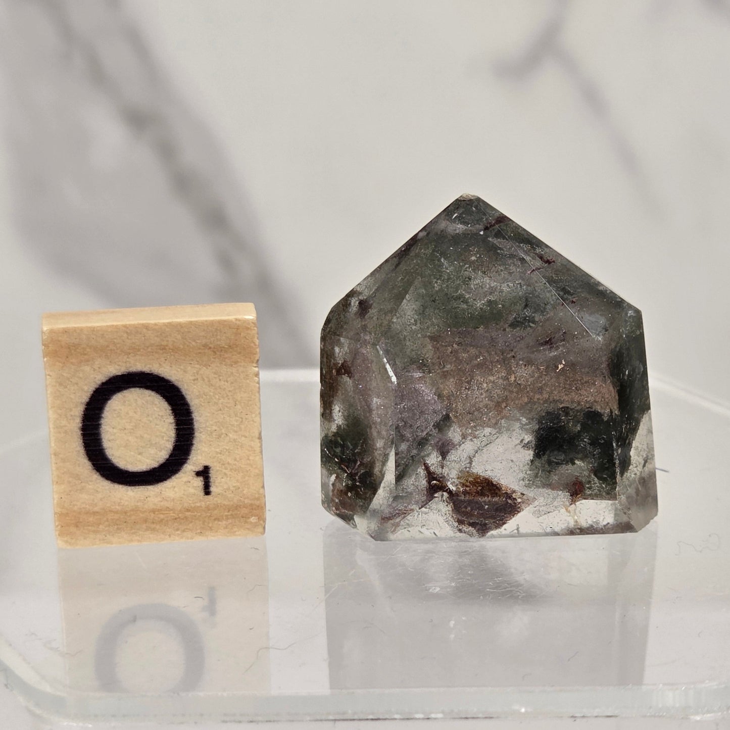 A small clear acrylic block with a black letter tile showing 'O1' next to a faceted natural crystal or stone specimen with dark gray, black, and white inclusions, displayed on a clear stand against a light marble-patterned background.