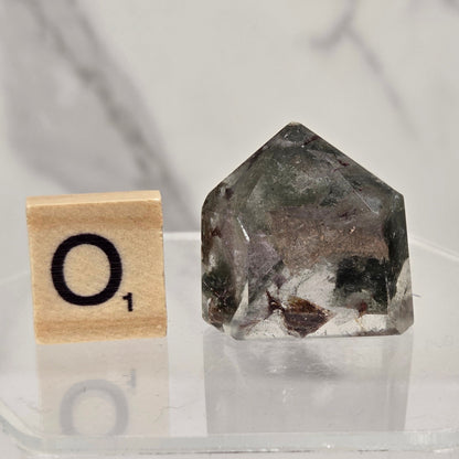 A small clear acrylic block with a black letter tile showing 'O1' next to a faceted natural crystal or stone specimen with dark gray, black, and white inclusions, displayed on a clear stand against a light marble-patterned background.