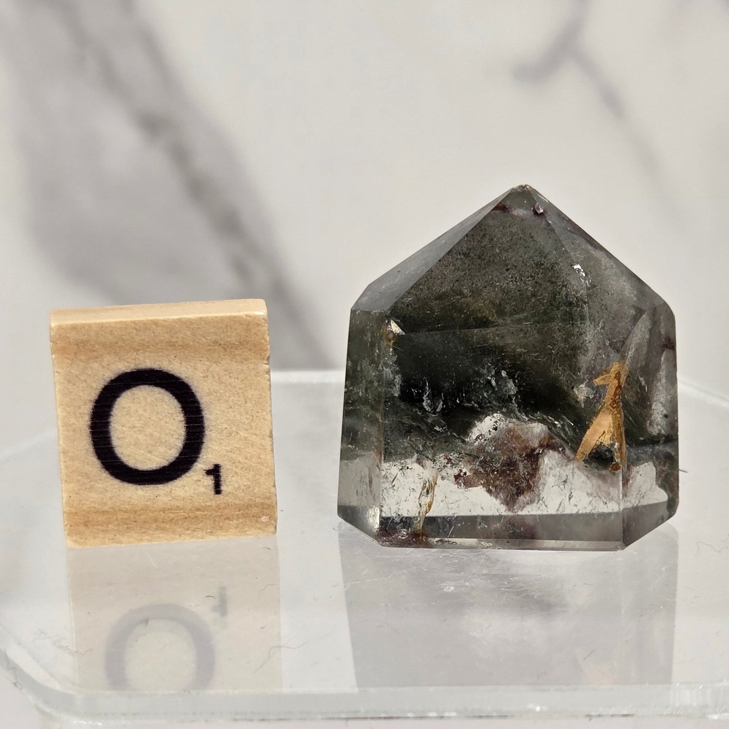 A clear faceted gemstone with dark inclusions and a small dried plant piece inside, displayed on a clear acrylic stand next to a wooden Scrabble tile showing the letter O.