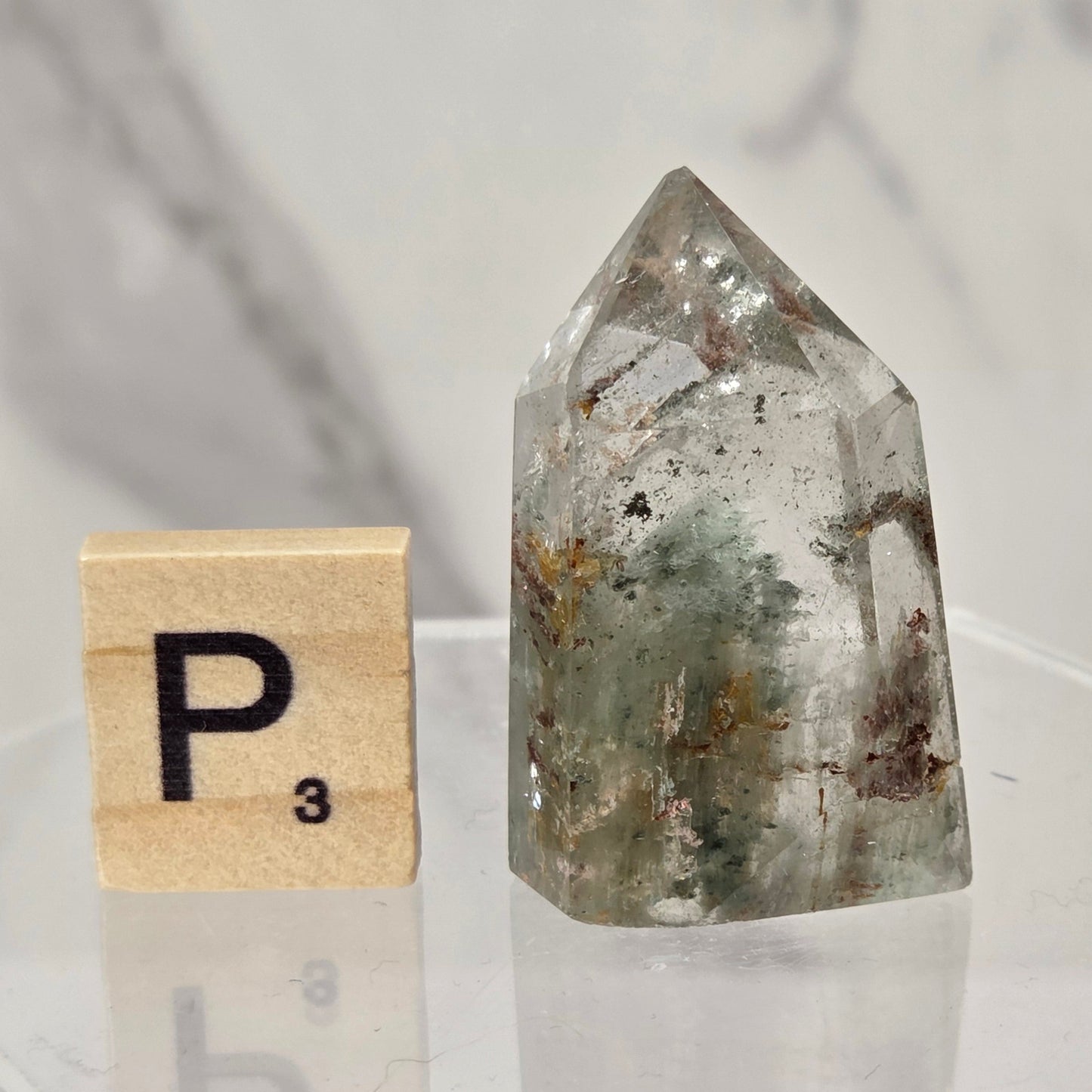 A pointed clear quartz crystal with internal green and brown inclusions, displayed next to a wooden Scrabble tile on a clear acrylic stand against a light marble background.
