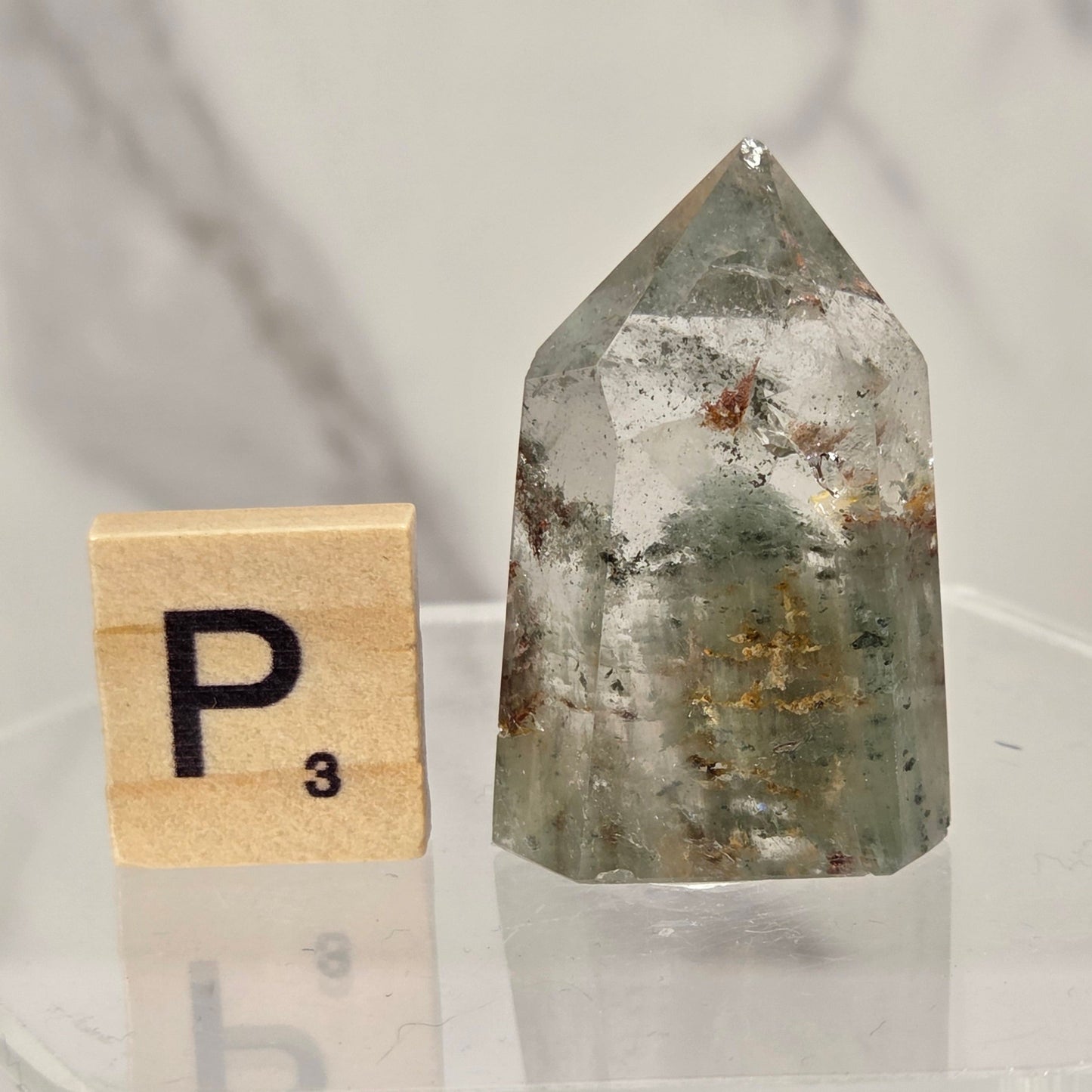 A pointed clear quartz crystal point with internal inclusions, displayed next to a wooden Scrabble tile on a clear acrylic stand against a marble background.