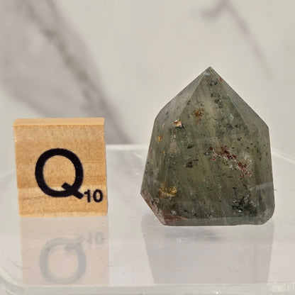 A faceted natural crystal point, likely a quartz crystal, with a greenish-gray body and small brown and reddish inclusions, displayed on a clear acrylic stand next to a wooden Scrabble tile showing the letter Q for 10 points.