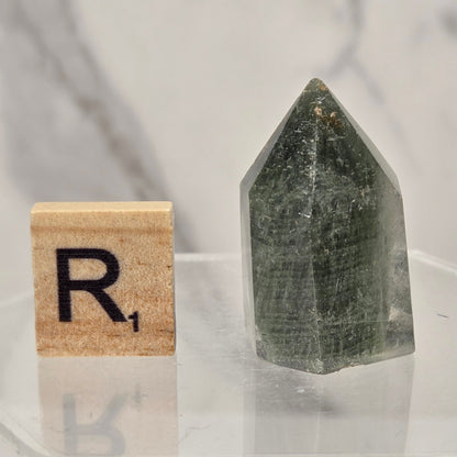 A pointed green crystal tower displayed next to a wooden Scrabble tile with the letter 'R' on a clear acrylic surface against a light marble background.