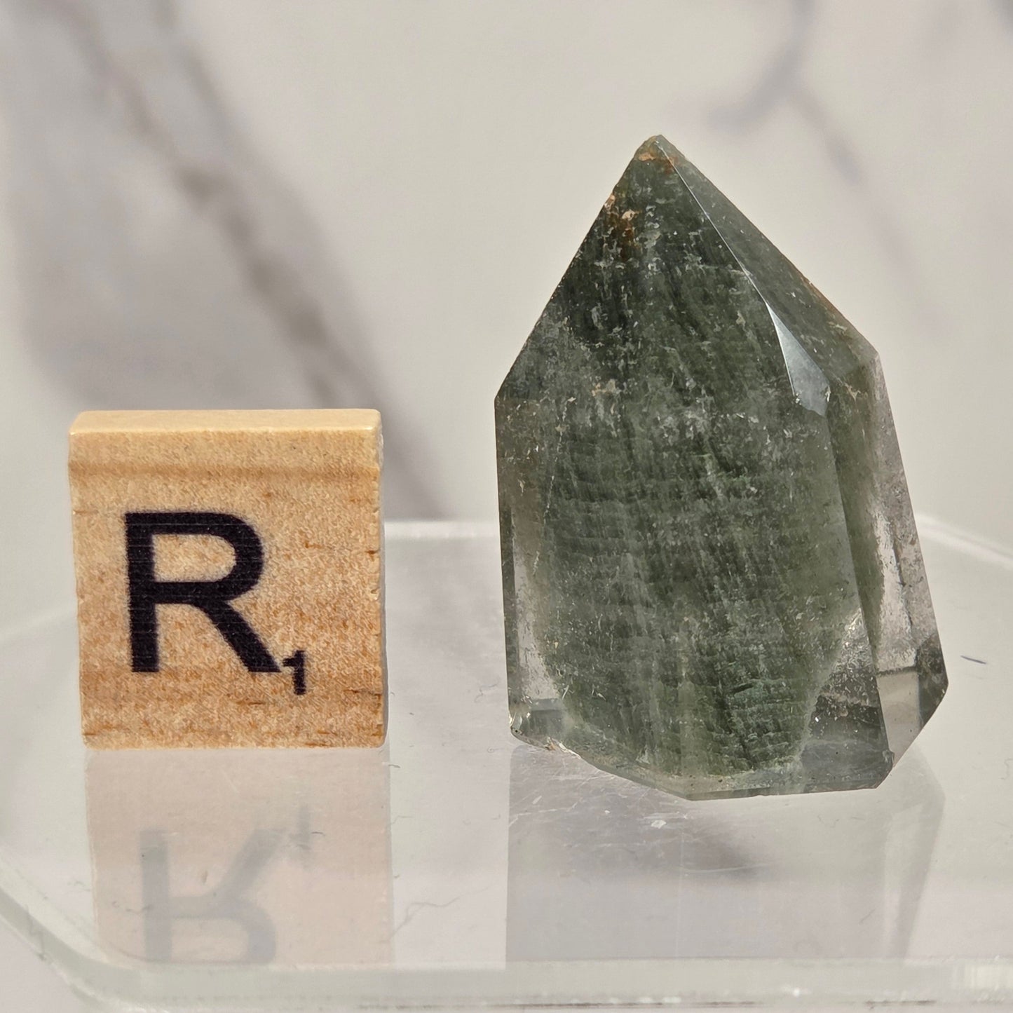A pointed clear quartz crystal with greenish-gray internal inclusions, displayed on a clear acrylic stand next to a wooden Scrabble tile showing the letter 'R'.