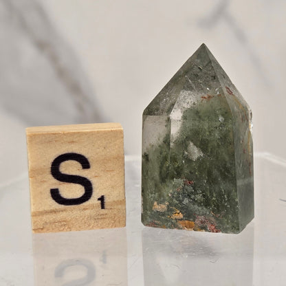 A pointed green crystal tower with internal inclusions sits on a clear acrylic block next to a wooden Scrabble tile showing the letter 'S'.