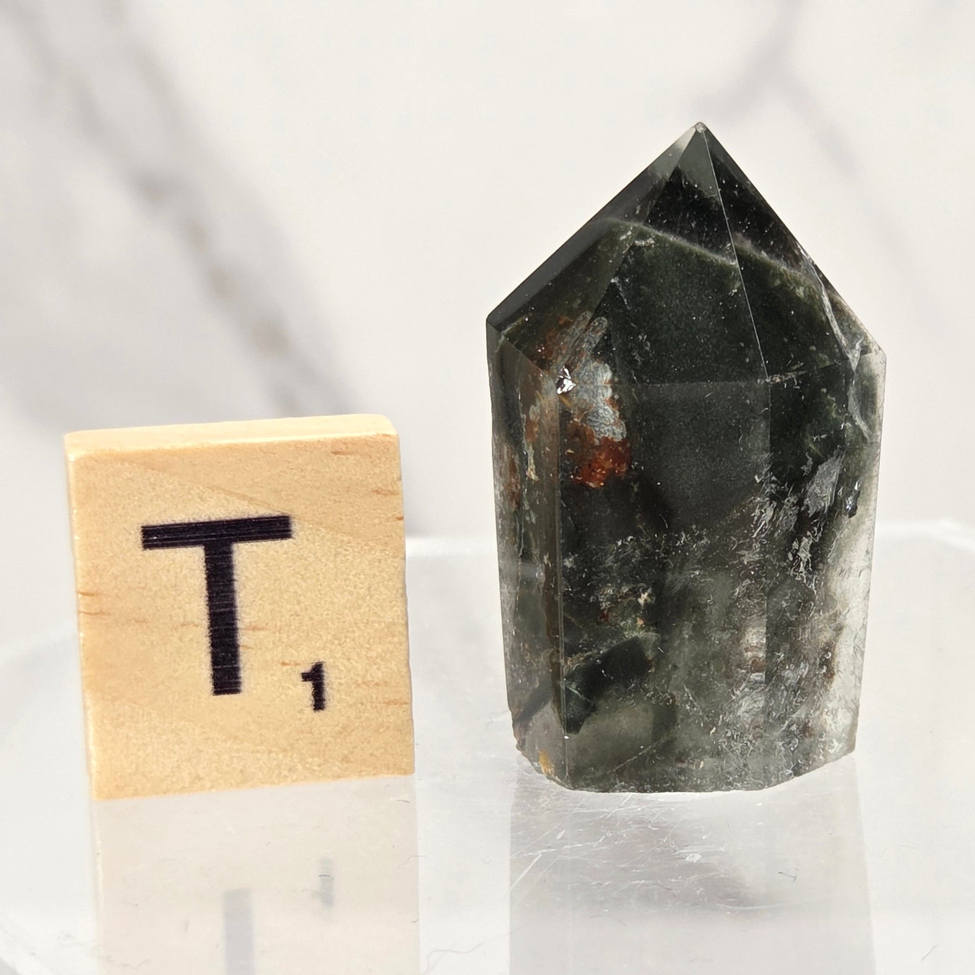 A dark green to black pointed crystal cluster or quartz point displayed on a clear acrylic stand next to a wooden Scrabble tile showing the letter T, against a light marble-patterned background.