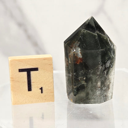 A dark green to black pointed crystal cluster or quartz point displayed on a clear acrylic stand next to a wooden Scrabble tile showing the letter T, against a light marble-patterned background.