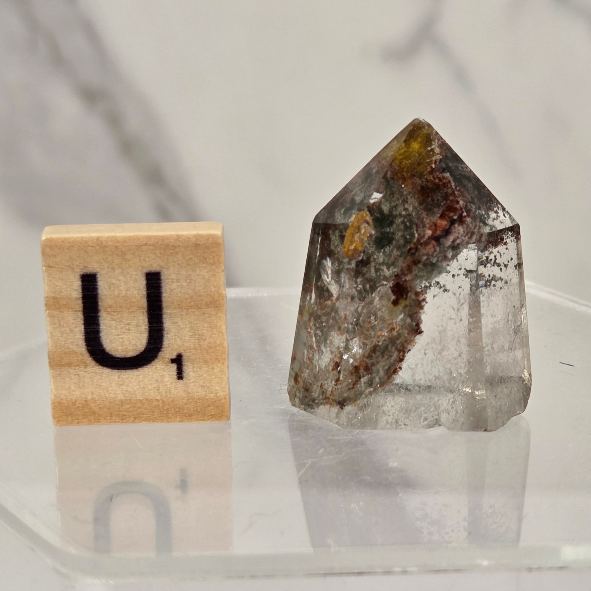 A clear pointed crystal point with internal brown, gray, and amber inclusions, displayed next to a wooden Scrabble tile marked 'U1' on a clear acrylic surface against a light marble background.
