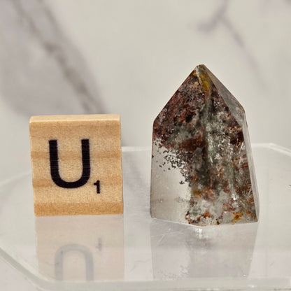 A pointed clear quartz crystal with brown and white inclusions, displayed next to a wooden Scrabble tile showing the letter 'U' on a clear acrylic stand.