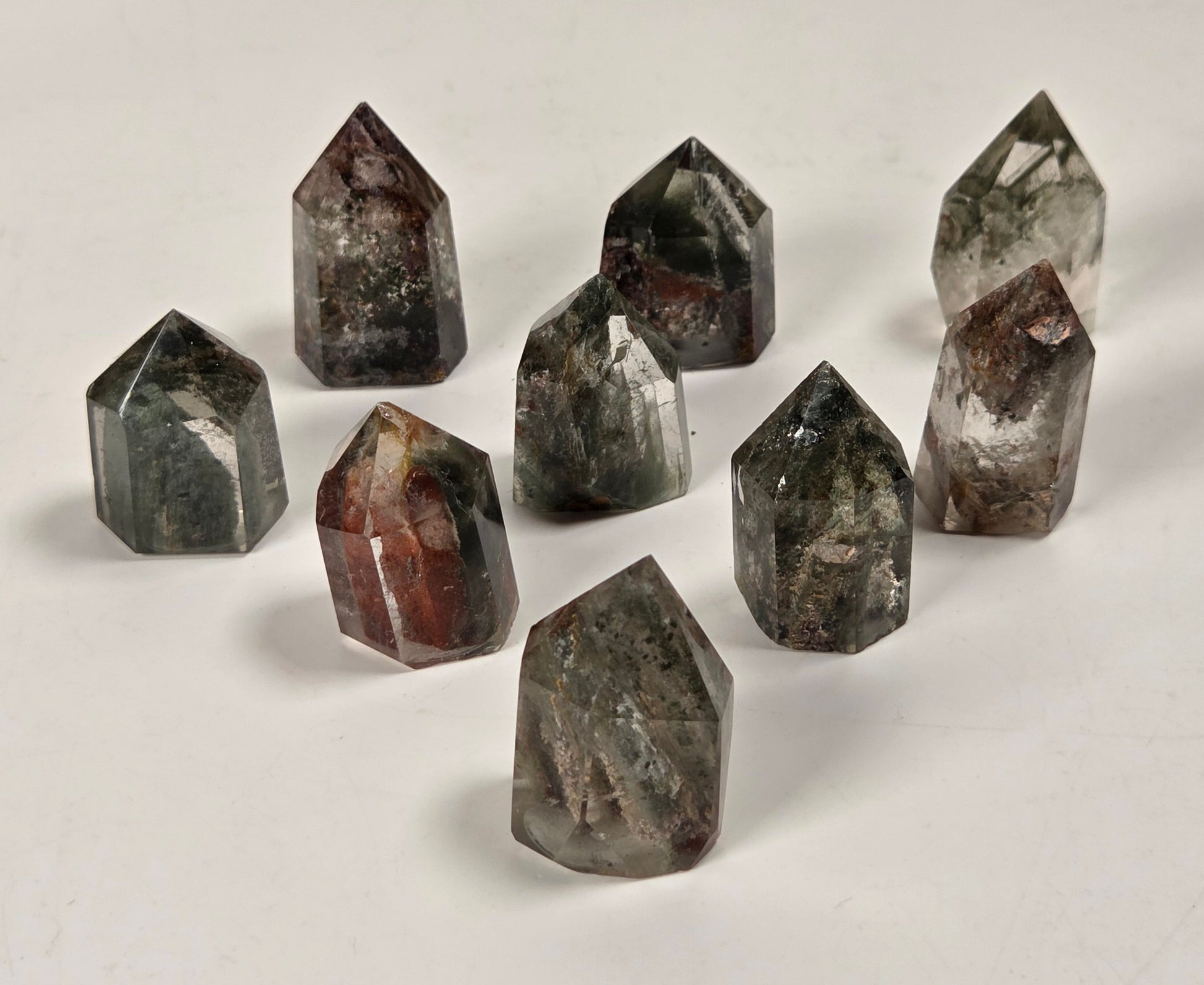 Nine polished crystal points of various shapes and colors, including gray, brown, and black with white inclusions, arranged on a plain off-white surface.