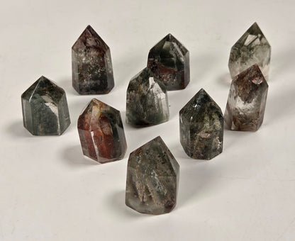 Nine polished crystal points of various shapes and colors, including gray, brown, and black with white inclusions, arranged on a plain off-white surface.