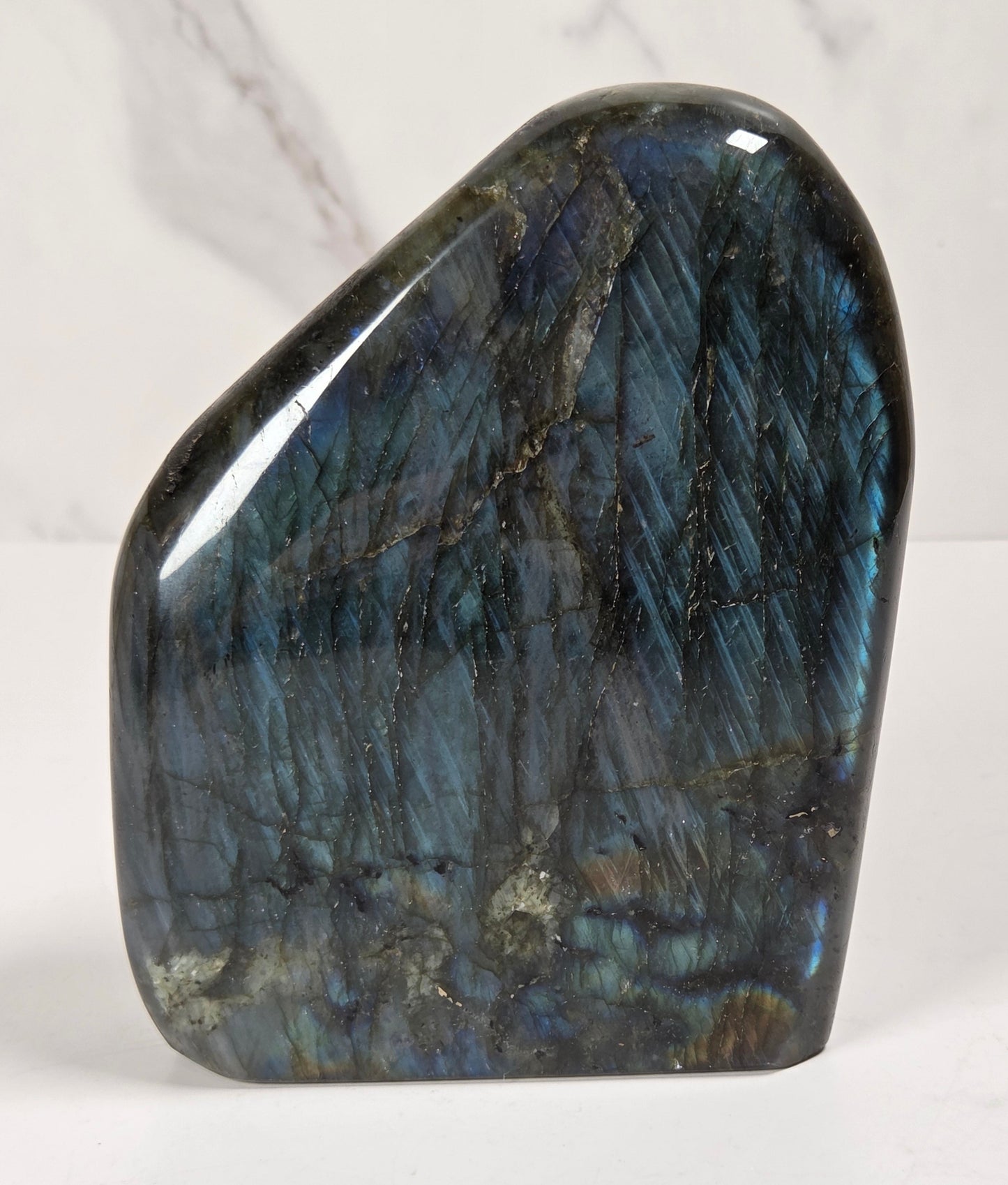 A polished labradorite crystal stone with a smooth, irregular shape, showcasing its characteristic blue and green iridescent flashes on a white surface with a light marble background.