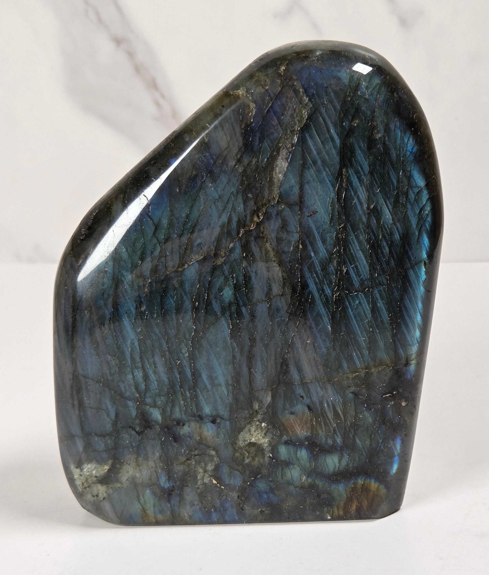 A polished labradorite crystal stone with a smooth, irregular shape, showcasing its characteristic blue and green iridescent flashes on a white surface with a light marble background.