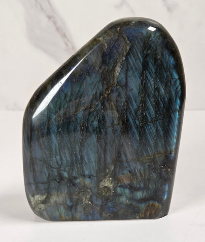 A polished labradorite crystal stone with a smooth, irregular shape, showcasing its characteristic blue and green iridescent flashes on a white surface with a light marble background.