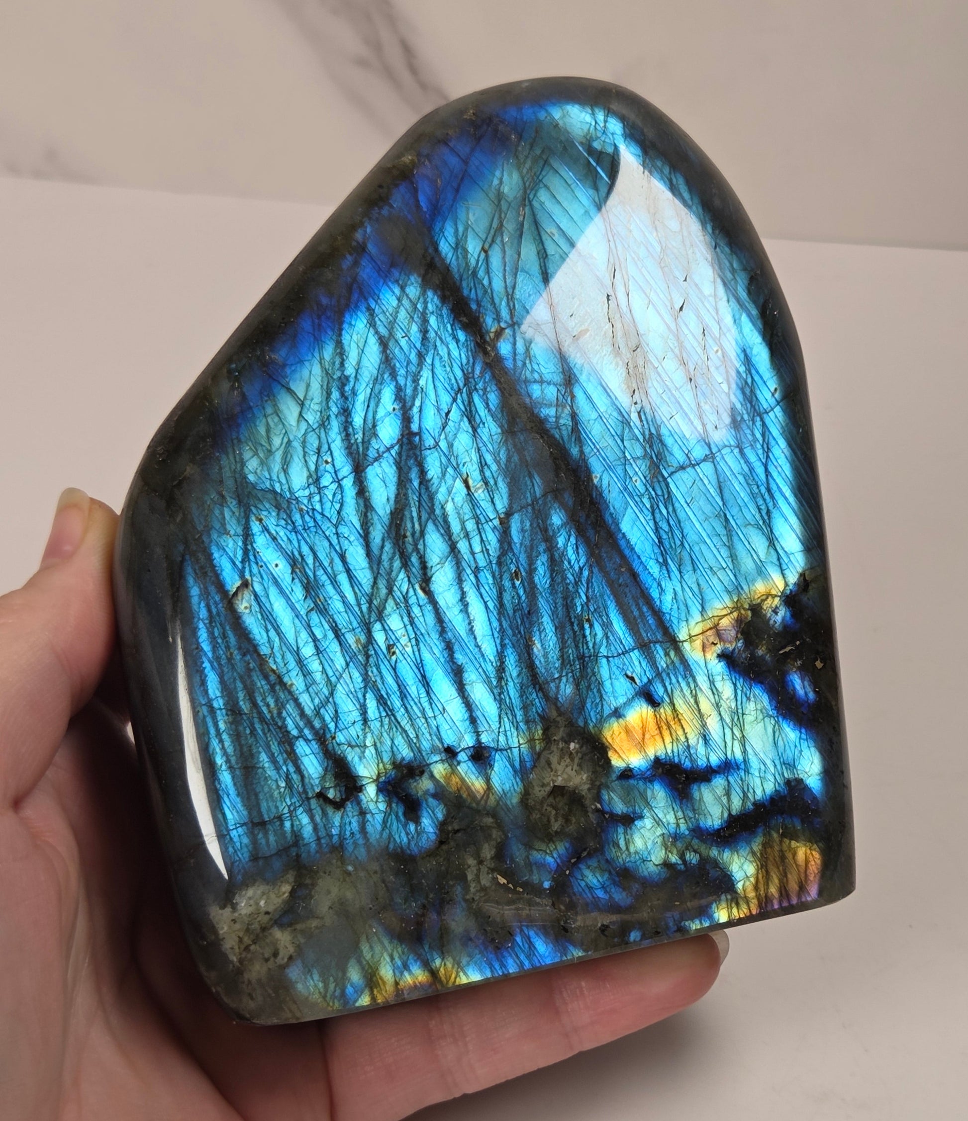 A hand holding a polished labradorite stone showing vibrant blue, green, and gold iridescent flashes, with dark inclusions throughout.