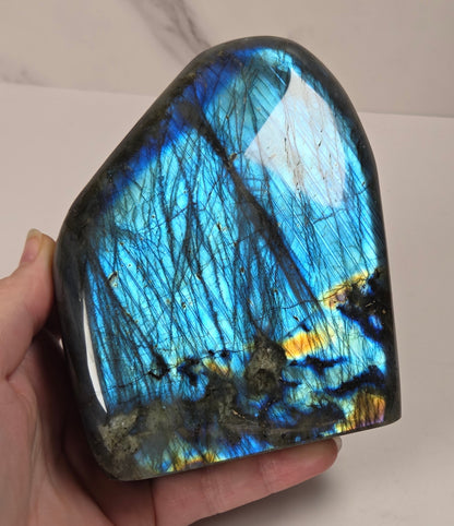 A hand holding a polished labradorite stone showing vibrant blue, green, and gold iridescent flashes, with dark inclusions throughout.