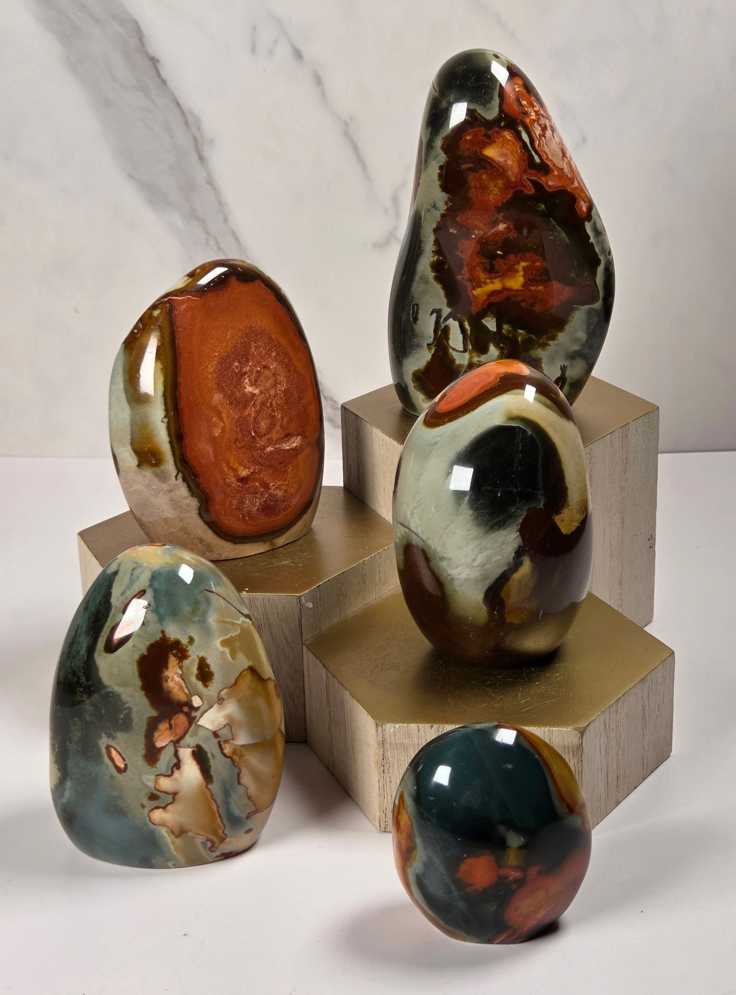 Five polished natural stone sculptures in various shapes and earthy color patterns, displayed on stacked gold-colored pedestals against a white marble-patterned background.