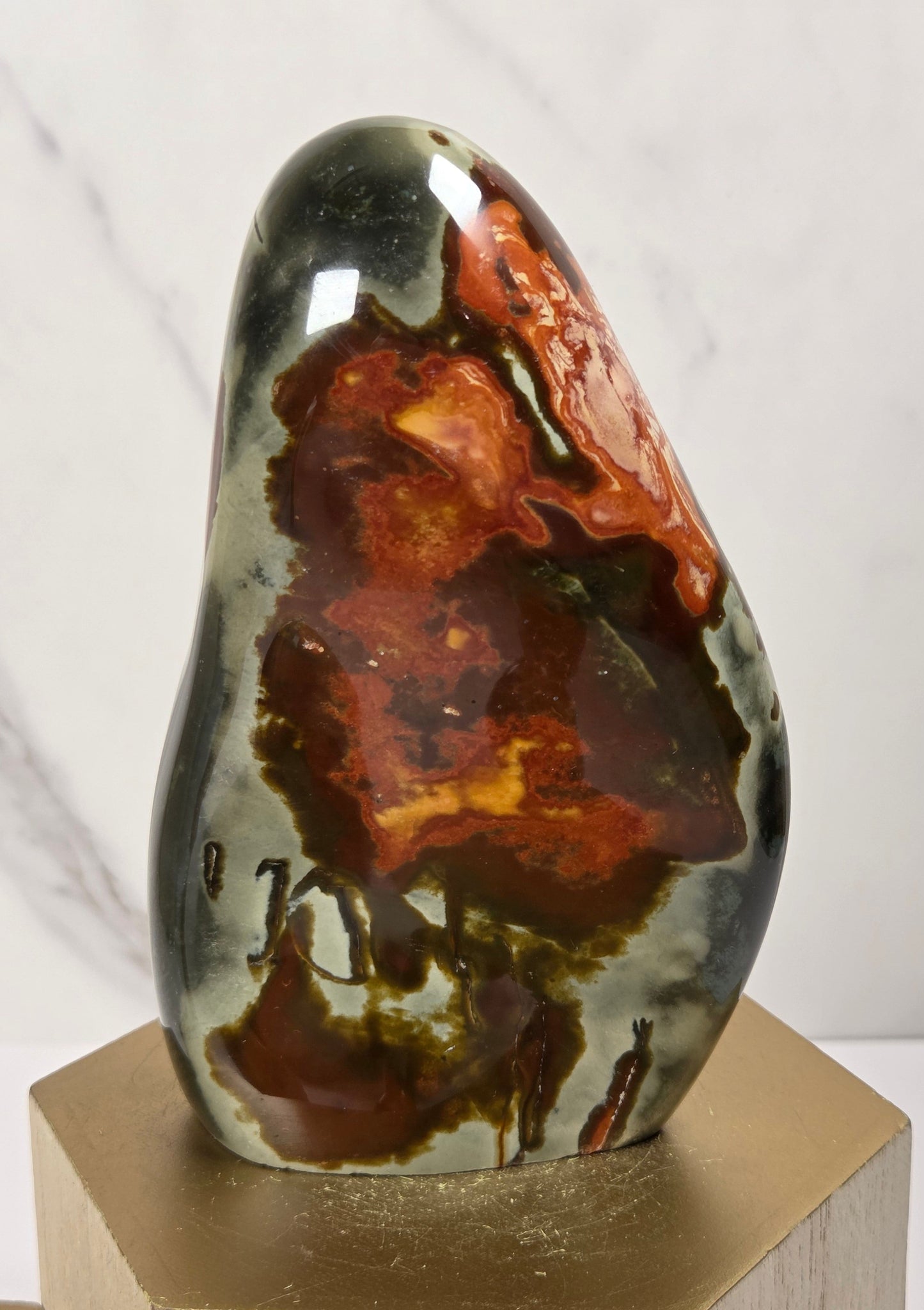 A polished natural stone sculpture with a glossy finish, featuring rich patterns of dark brown, rust red, and black over a gray-green base, displayed on a hexagonal gold-toned wooden pedestal against a white marble background.