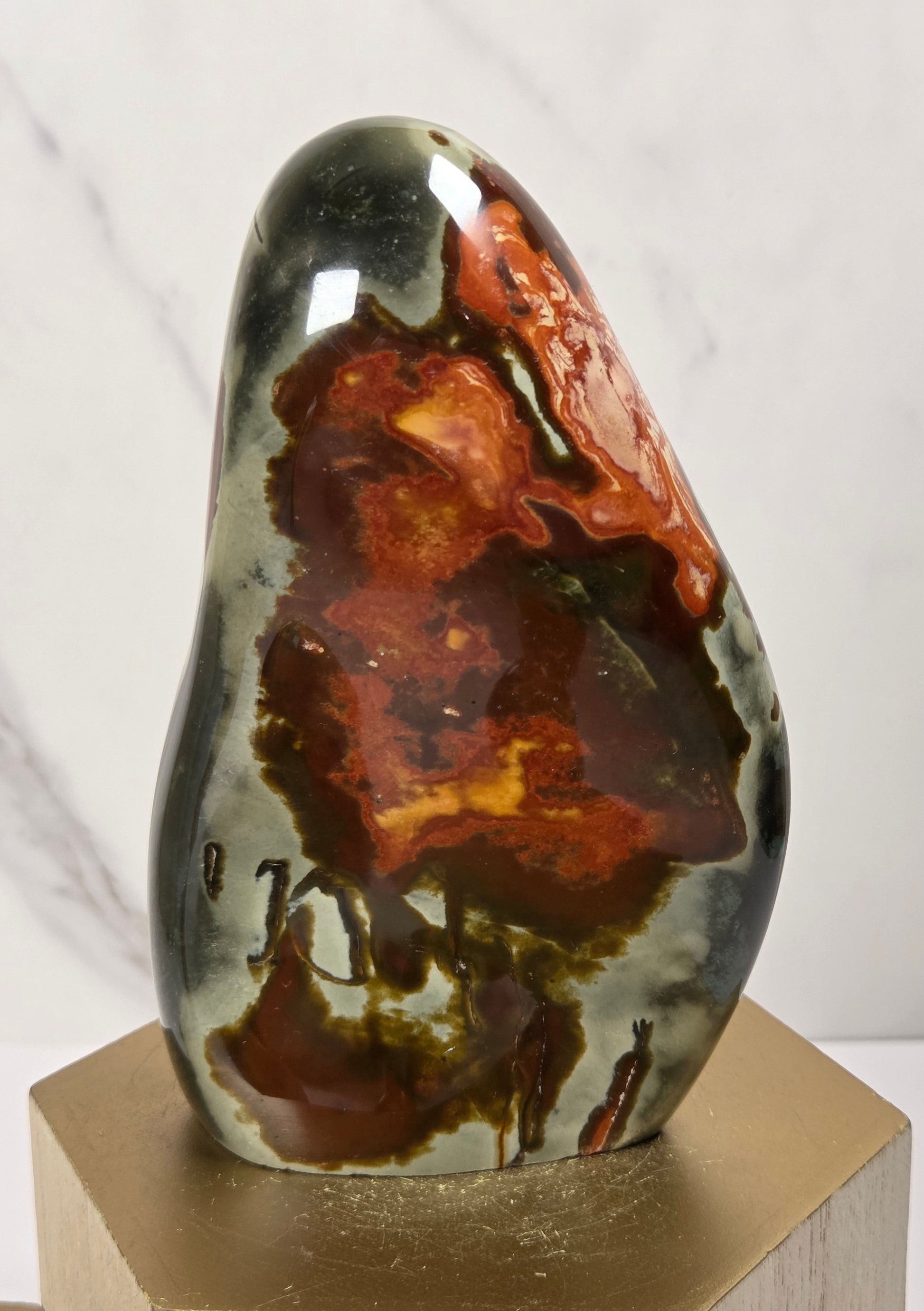 A polished natural stone sculpture with a glossy finish, featuring rich patterns of dark brown, rust red, and black over a gray-green base, displayed on a hexagonal gold-toned wooden pedestal against a white marble background.