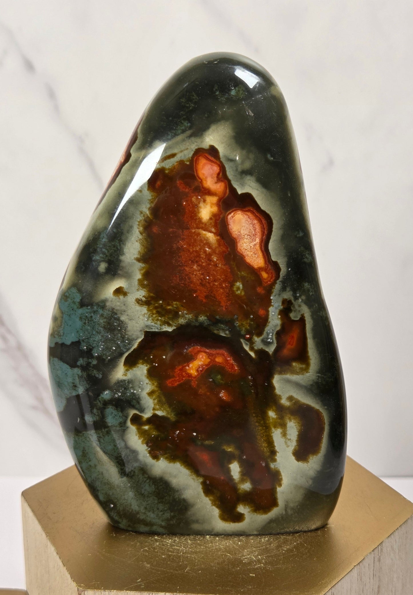 A polished natural stone sculpture with a dark green, gray, and brown agate-like pattern, displayed on a gold hexagonal pedestal against a light marble background.