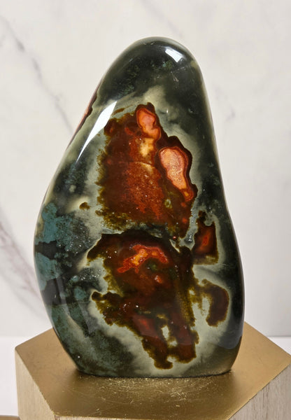 A polished natural stone sculpture with a dark green, gray, and brown agate-like pattern, displayed on a gold hexagonal pedestal against a light marble background.