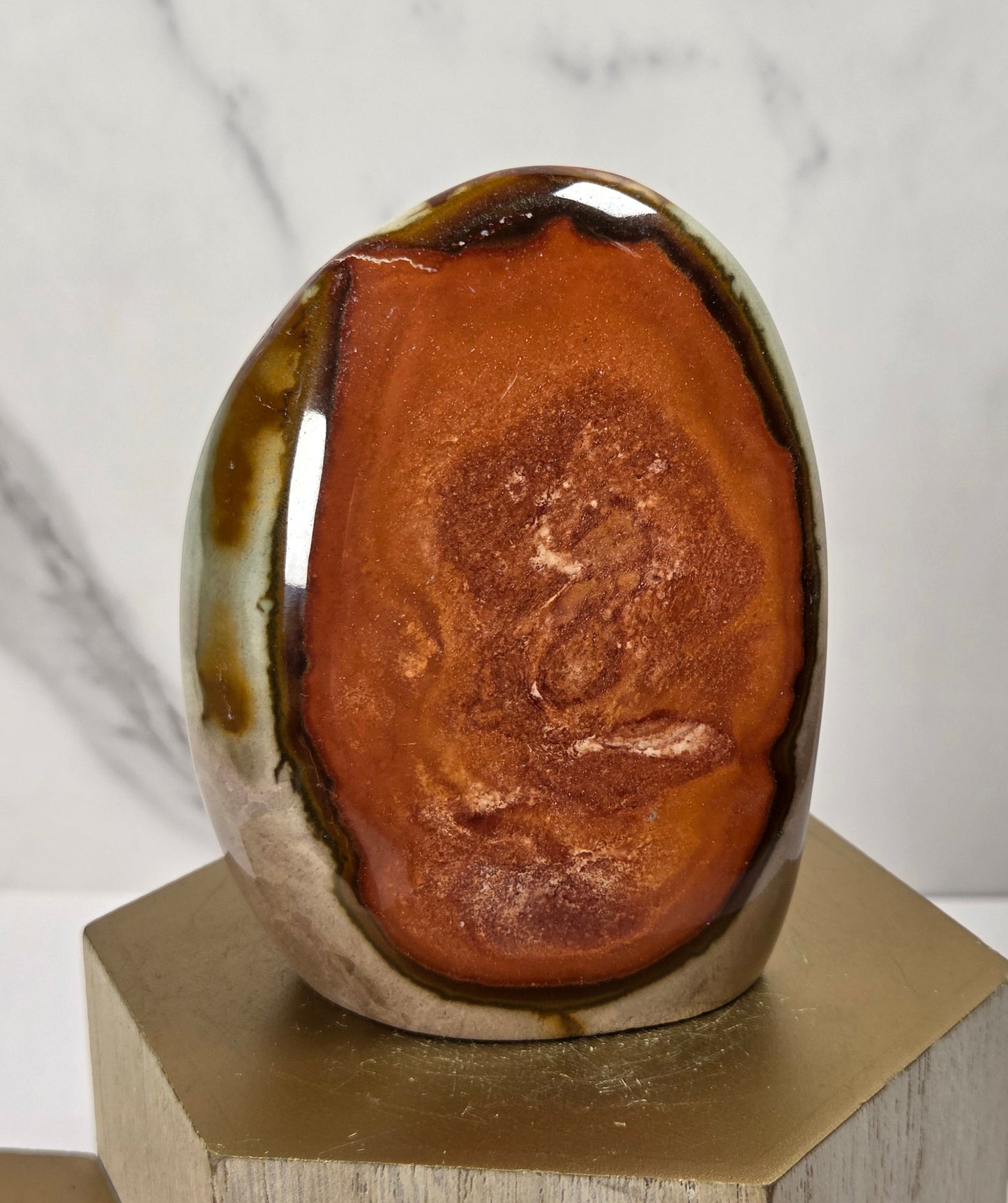 A polished ceramic egg-shaped sculpture with a glossy brown and amber center and a glazed green and beige outer edge, displayed on a hexagonal wooden pedestal against a light marble-patterned background.