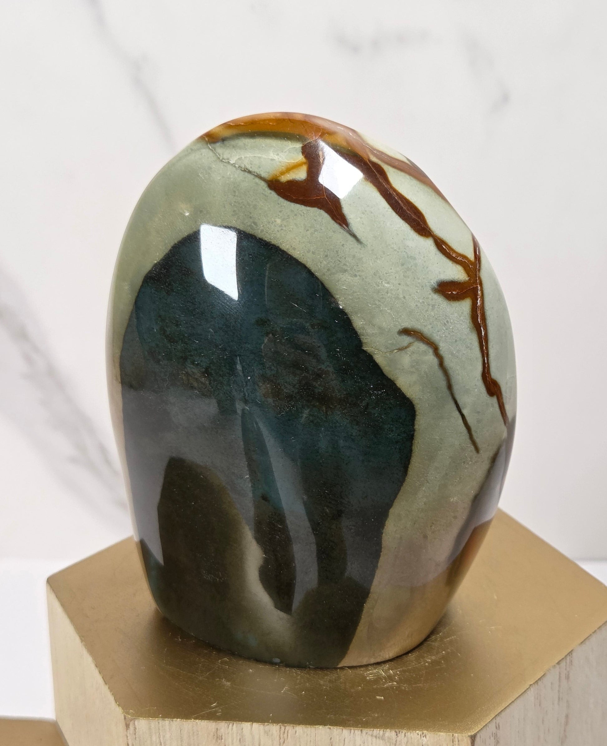 A polished stone sculpture with a smooth, rounded organic shape, featuring a marbled pattern of dark teal, olive green, cream, and brown veins, displayed on a gold hexagonal pedestal against a white marble background.