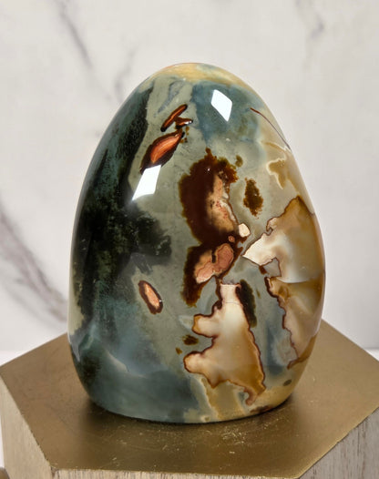 A polished agate stone egg sculpture with natural marbled patterns in green, brown, and beige, displayed on a wooden block pedestal against a marble-patterned background.