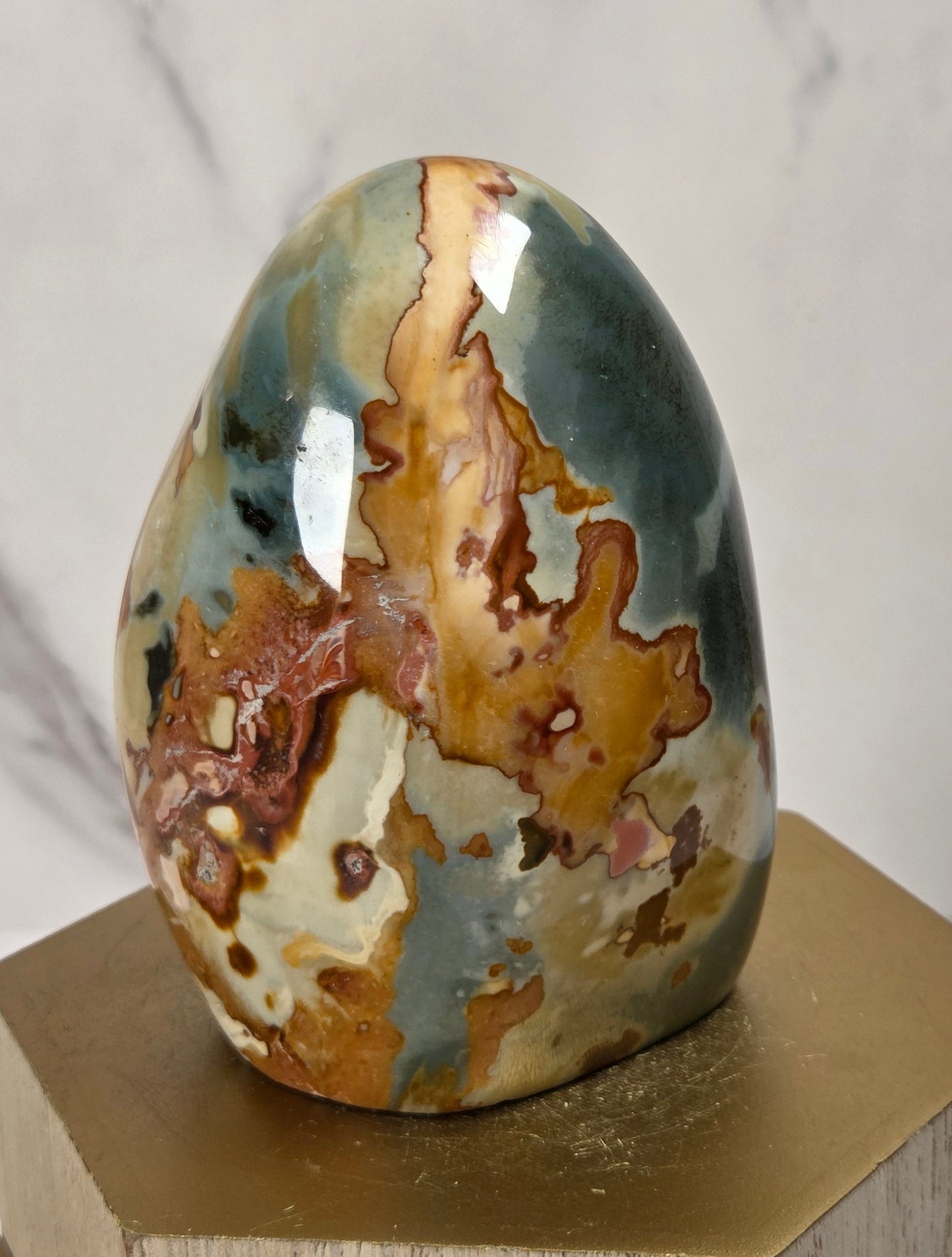 A polished natural stone egg sculpture with mottled earthy tones of brown, beige, gray, and muted blue-green, displayed on a hexagonal gold-toned pedestal against a light marble-patterned background.
