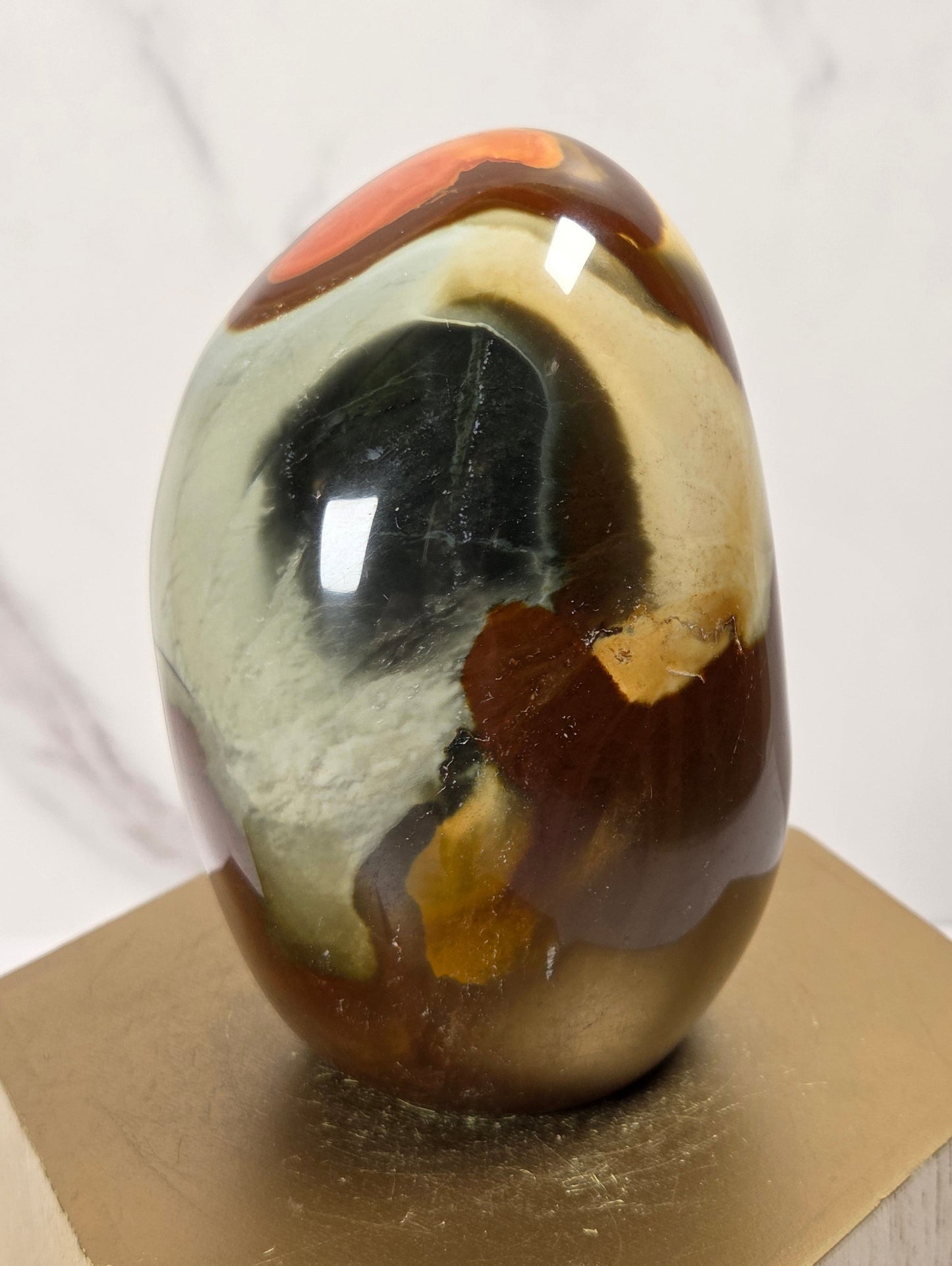A polished agate stone egg with marbled patterns of brown, cream, black, and orange, displayed on a gold hexagonal stand against a light marble background.