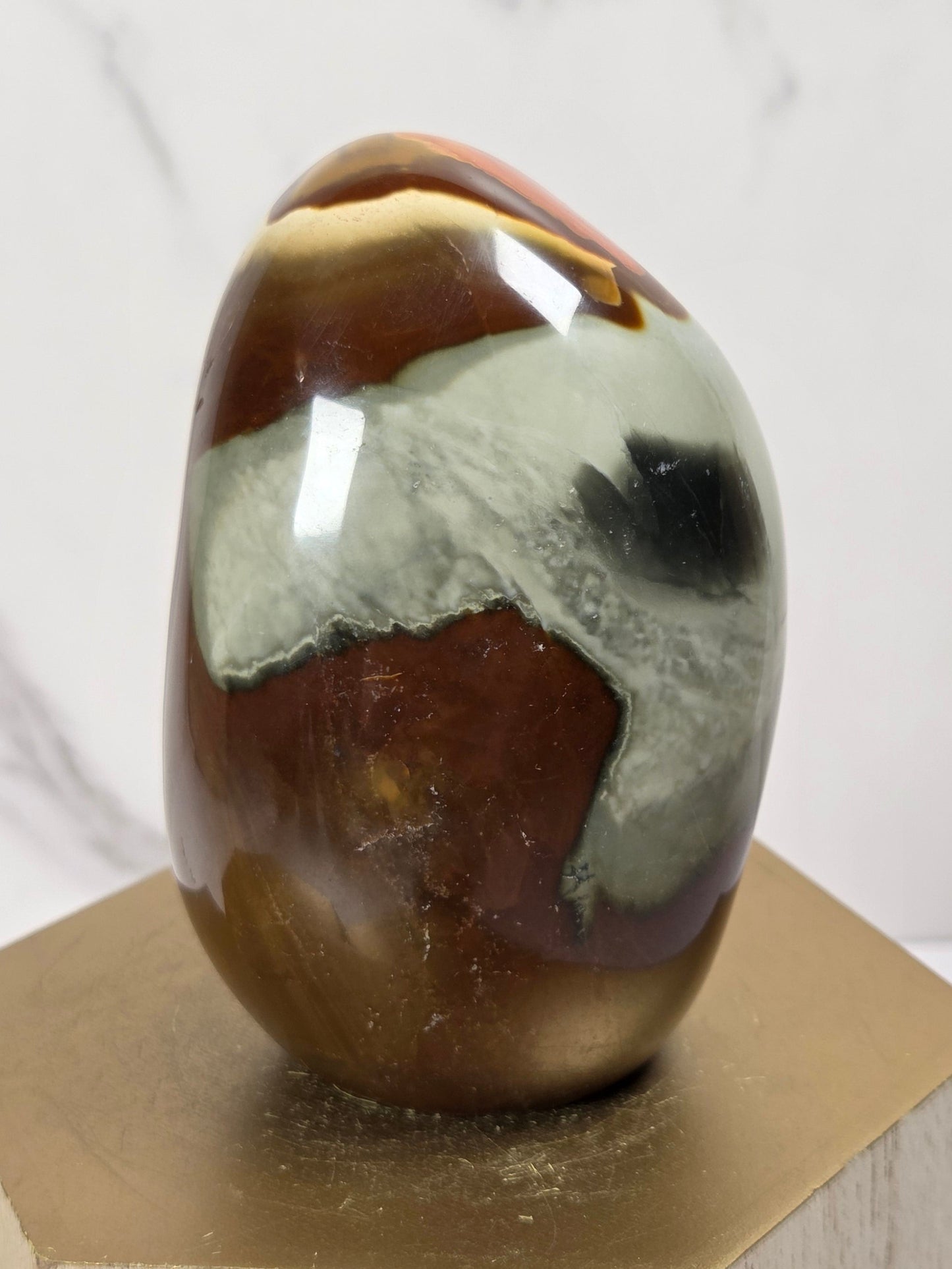 A polished oval stone sculpture with marbled brown, cream, gray, and black patterns, displayed on a hexagonal gold-toned pedestal against a light marble-patterned background.