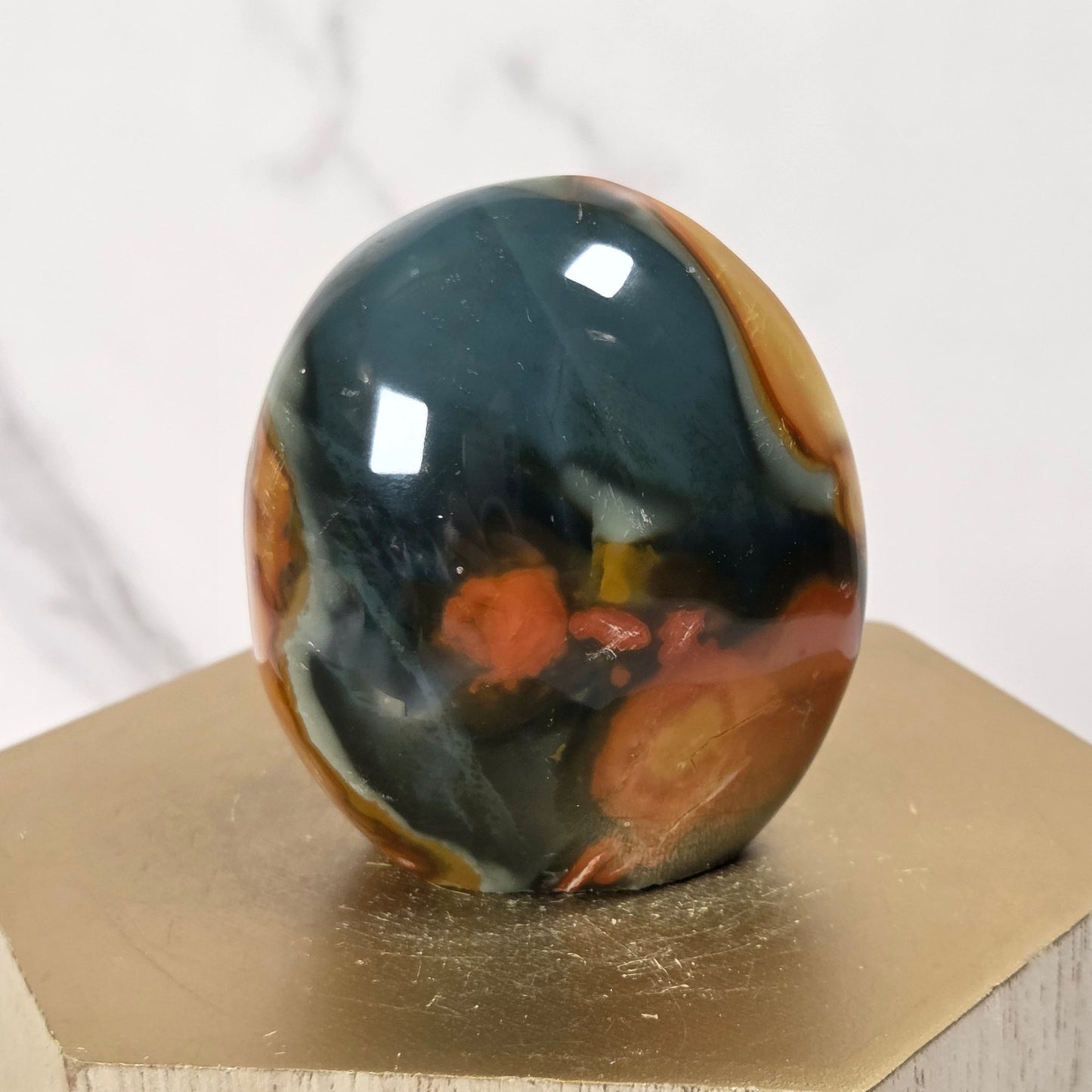A polished oval stone with a marbled pattern of dark gray, black, orange, and brown sits on a gold hexagonal display block against a light marble background.