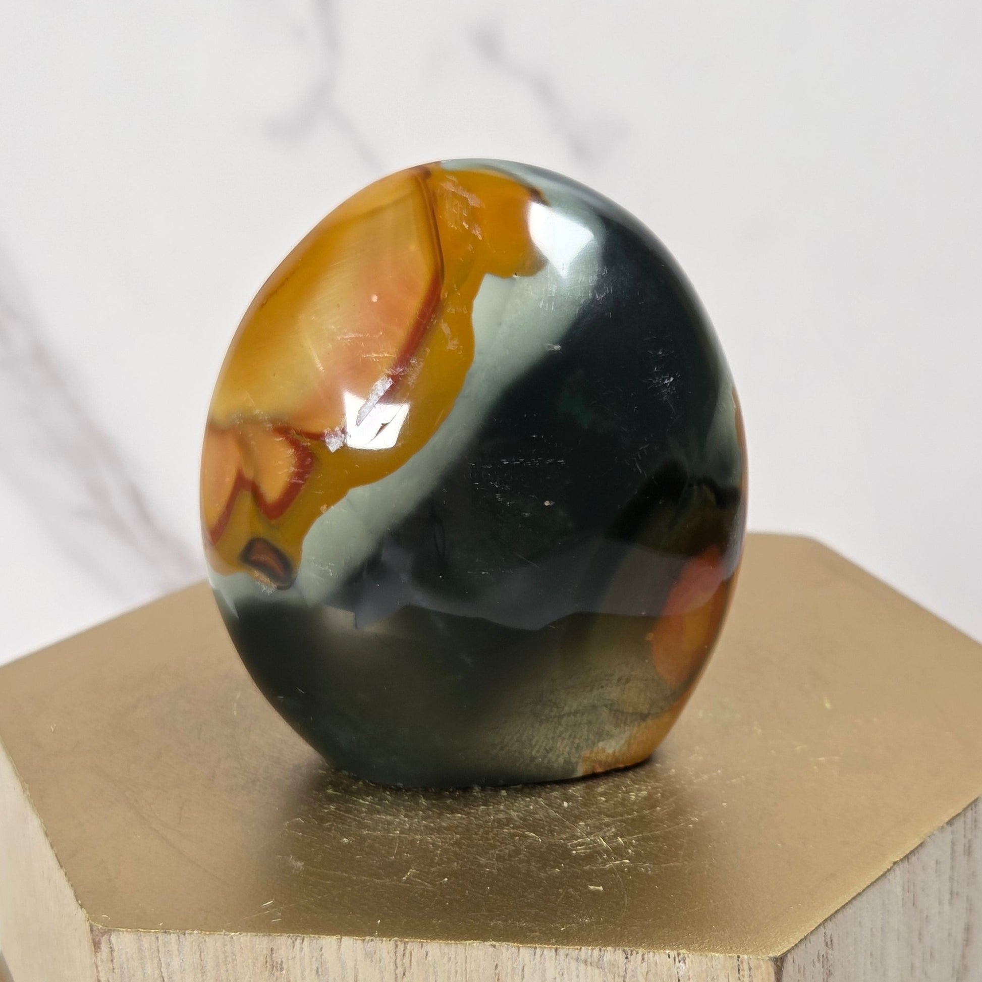 A polished agate stone egg with swirling patterns of black, orange, and cream, displayed on a hexagonal gold-colored pedestal against a white marble background.