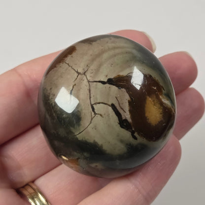 A hand holding a polished round stone with a mottled pattern of brown, beige, and dark gray, featuring a glossy finish and natural fissures.