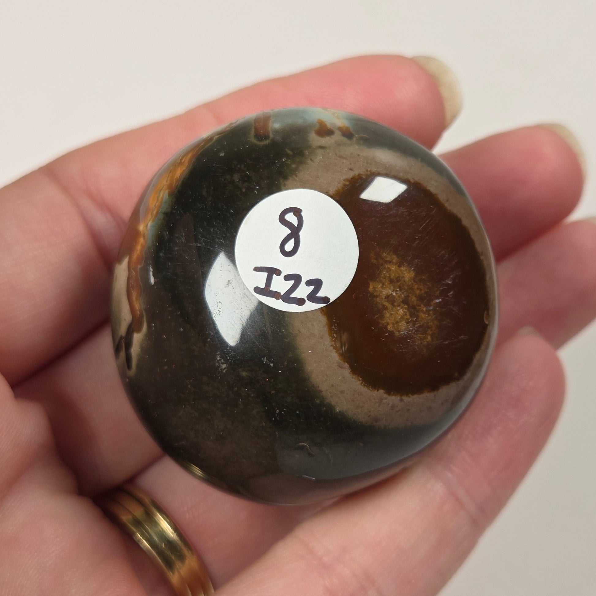 A hand holding a polished round agate stone with natural banding in dark brown, black, and tan, featuring a small white sticker with the number 8122 on the front.