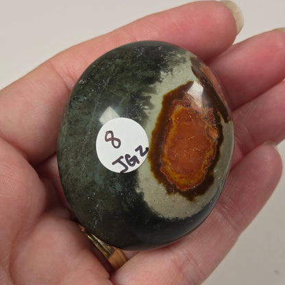 A hand holding a polished oval stone with a dark green and black outer area, a cream band, and a prominent orange-brown center, with a small white sticker labeled '8 JG2' on the surface.