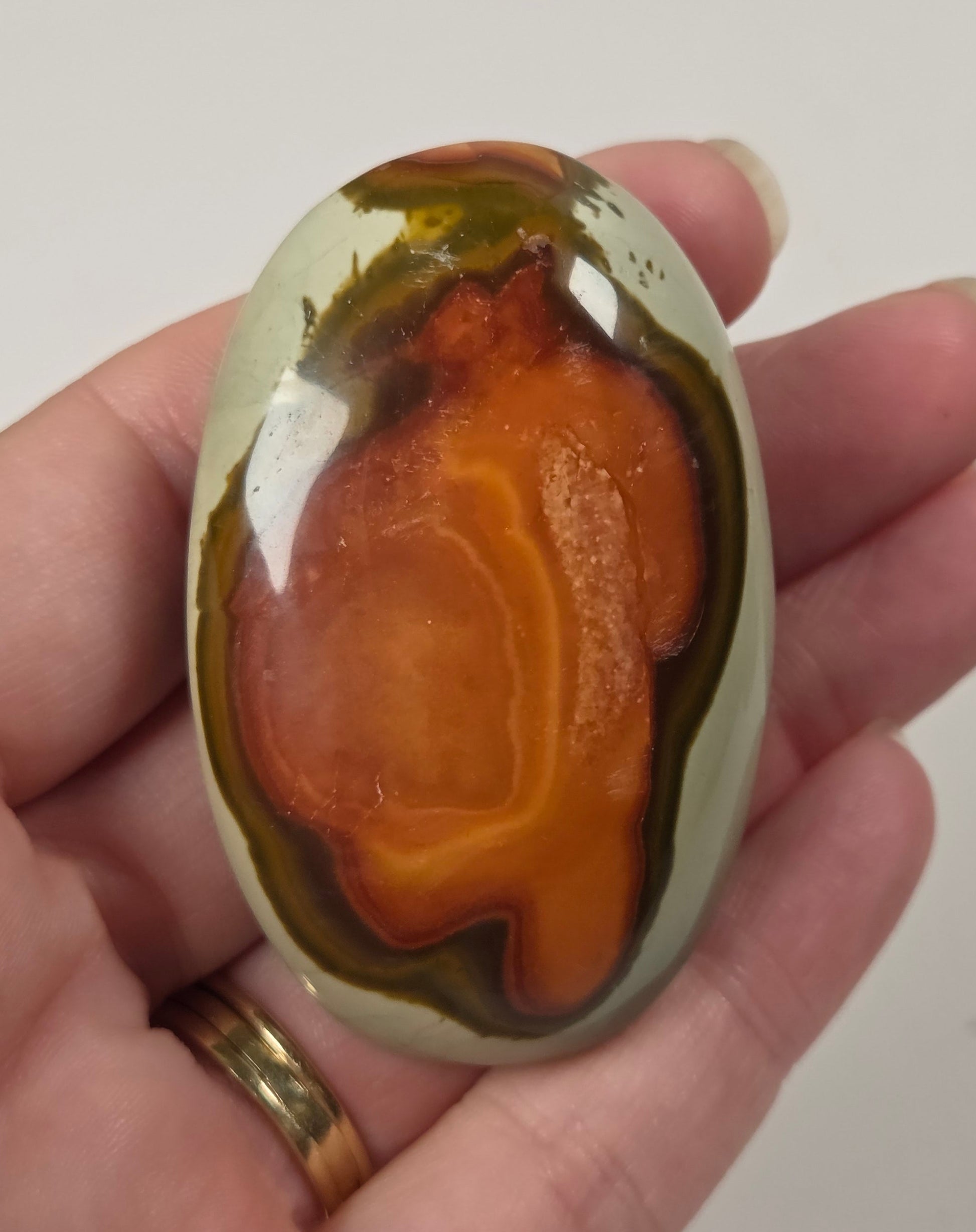 A hand holding a polished oval agate stone with concentric bands of orange, brown, olive green, and cream.