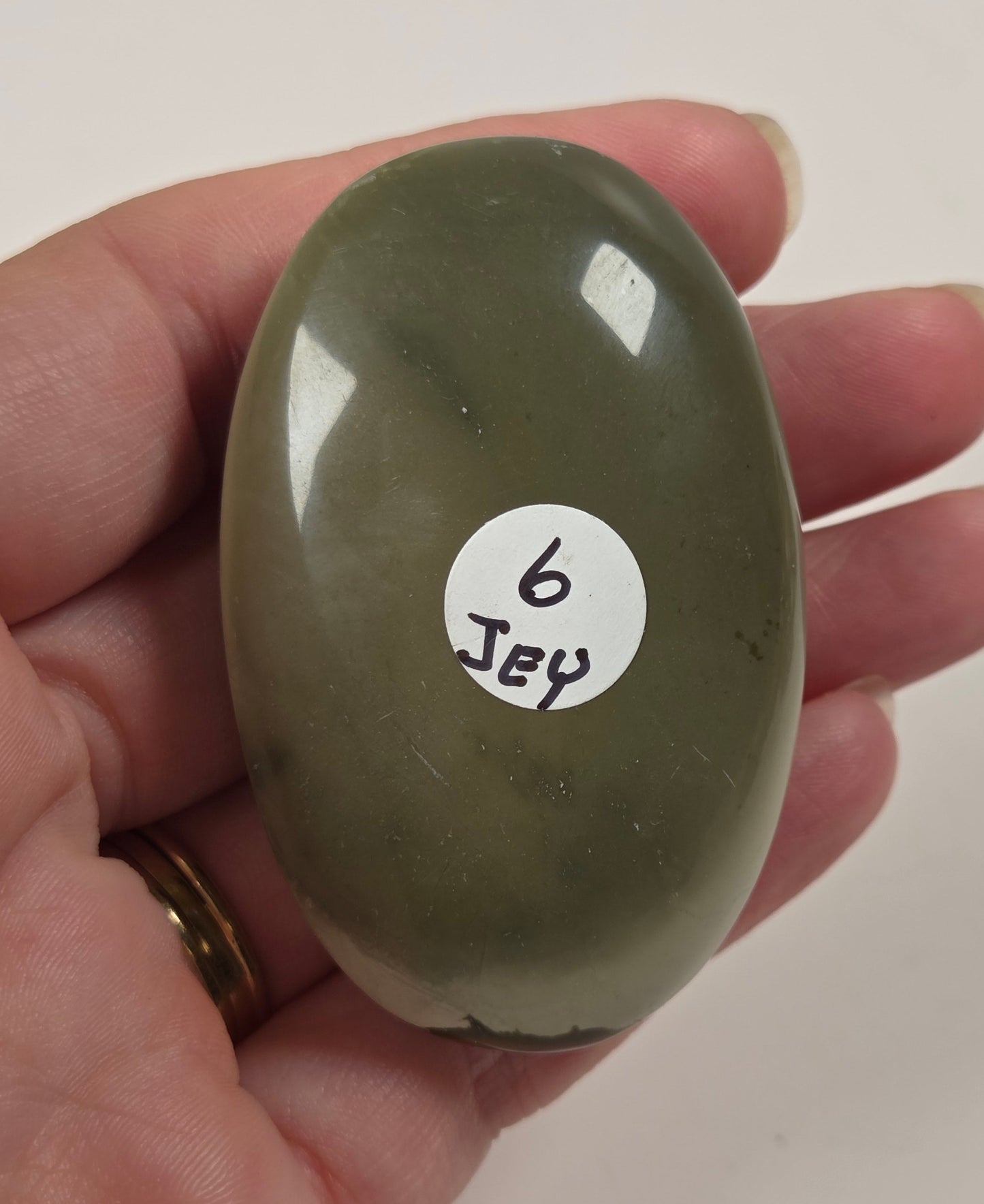 A hand holding a smooth, polished olive green stone with a small white sticker that reads '6 Jey' on its surface, set against a plain off-white background.