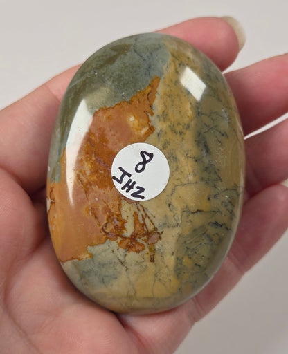 A hand holding a polished, oval-shaped stone with natural mottled patterns of olive green, golden yellow, and orange-brown. A small white sticker with handwritten text is attached to the stone.